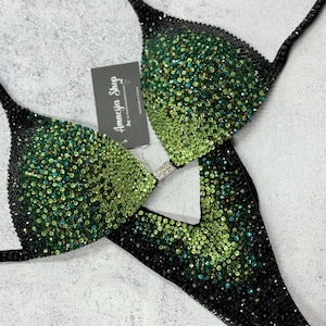 Puede incluir: Bikini negro y verde con adornos de cristal. La parte superior del bikini tiene una tira de estilo halter y la parte inferior es de estilo de corte alto. El bikini está decorado con cristales verdes y negros.