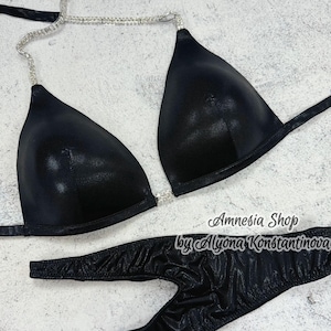 Puede incluir: Bikini negro de arriba y abajo con una tira de cadena de cristal. La parte superior tiene forma de triángulo y la parte inferior es de estilo de corte alto. El texto "Amnesia Shop by Alyona Konstantinova" está impreso en la parte inferior.