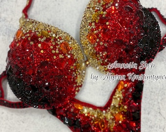 May include: A red, black, and gold sequined competition swimsuit with a V-neckline and a high-cut leg. The swimsuit is decorated with rhinestones and crystals. "Amnesia Shop by Alyona Konstantynova" is written on the swimsuit.
