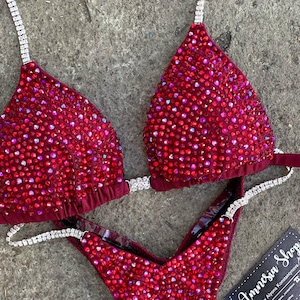 Puede incluir: Un traje de competición de bikini rojo y plateado con lentejuelas y pedrería. El traje tiene una parte superior triangular y una parte inferior en forma de V. El traje está sobre un fondo gris. El texto "Amnesia Shop" está en una tarjeta negra en la esquina inferior derecha de la imagen.