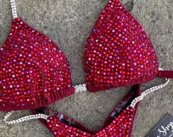 May include: A red and silver sequined bikini competition suit with rhinestones. The suit has a triangle top and a V-shaped bottom. The suit is on a gray background. The text "Amnesia Shop" is on a black card in the bottom right corner of the image.