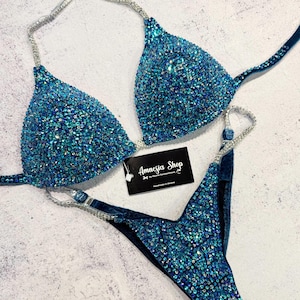 Op de afbeelding: Een turquoise bikiniset met sprankelende versieringen. De top heeft driehoekige cups met strass bandjes en de onderkant is een V-vormig ontwerp. Het label luidt "Amnesia Shop".