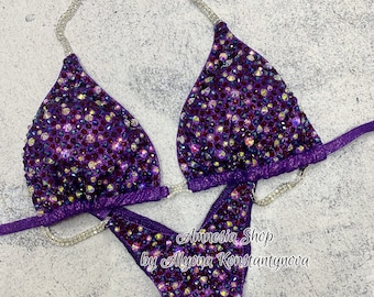 May include: Purple and silver sequined bikini competition suit with rhinestones. The suit has a halter top and a bottom with a high cut leg. The text "Amnesia Shop by Alyona Konstantynova" is printed on the bottom.
