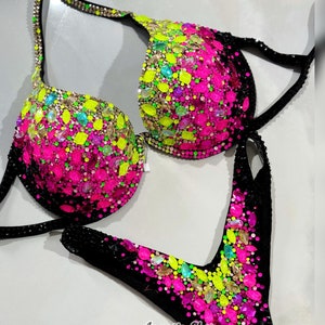 May include: A black bikini with a pink and green ombre design. The bikini is covered in rhinestones and crystals. The text "Amnesia Shop by Alyona Konstantinova" is printed on the bottom of the bikini.