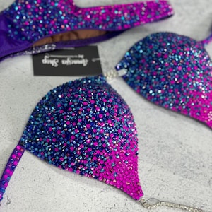Puede incluir: Un top de bikini morado y rosa con un degradado de pedrería. El top está decorado con pedrería azul, rosa y morada.