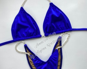 Puede incluir: Un bikini de competición azul real con la parte inferior en forma de V y la parte superior triangular. El bikini está adornado con tirantes con incrustaciones de cristal. El texto "Amnesia Shop by Alyona Konstantinova" es visible en la imagen.