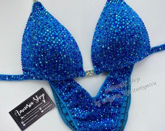 May include: A turquoise blue competition bikini with crystal embellishments. The bikini has a halter top and a high-cut bottom. The bikini is decorated with rhinestones and crystals. The bikini is made of a stretchy fabric. The bikini is designed for competition and is very sparkly.