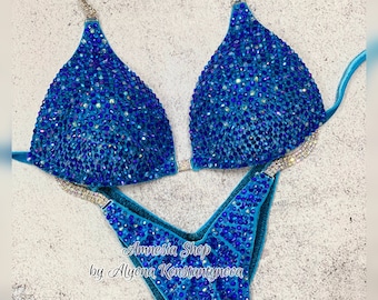 May include: A turquoise blue bikini competition suit with crystal embellishments. The suit has a halter top and a V-shaped bottom. The text "Amnesia Shop by Alyona Konstantynova" is printed on the bottom.