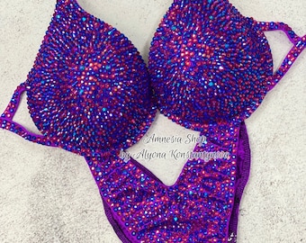 May include: Purple and blue jeweled competition bikini with a V-neck and a high-cut leg. The bikini is covered in rhinestones and has a shiny finish. The text "Amnesia Shop by Alyona Konstantynova" is printed on the bottom of the bikini.