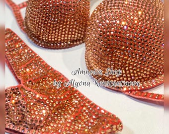 Op de afbeelding: Een koraalkleurige bikini top en broek met een hoogglans afwerking. De stof is bedekt met kleine, glinsterende strass steentjes. De bikini is gelabeld "Amnesia Shop by Alyona Konstantinova."