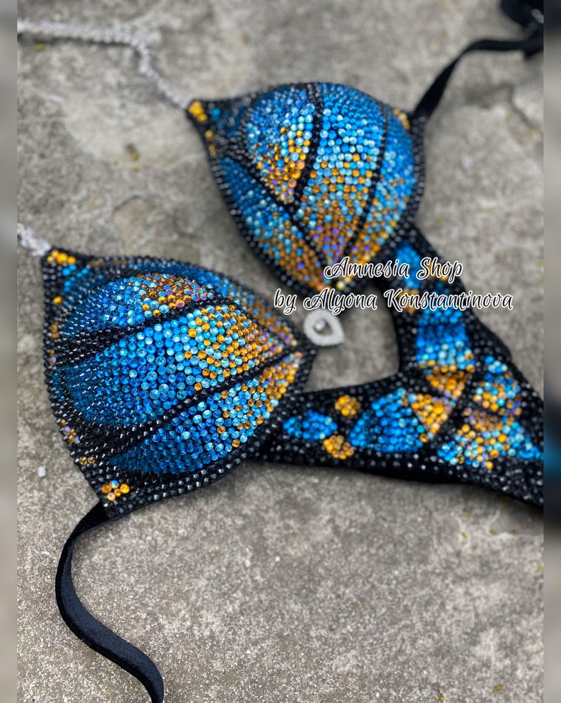 Op de afbeelding: Een zwarte bikini top en broek met blauwe en gouden steentjes. De bikini is versierd met een geometrisch patroon. De tekst "Amnesia Shop by Alyona Konstantinova" is zichtbaar op de afbeelding.