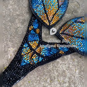 Op de afbeelding: Een zwarte bikini met blauwe en gouden steentjes. De bikini top heeft een schelp ontwerp en de onderkant heeft een vlinder ontwerp. De bikini is versierd met de tekst "Amnesia Shop by Alyona Konstantinova".