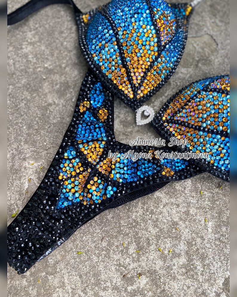 Op de afbeelding: Een zwarte bikini met blauwe en gouden steentjes. De bikini top heeft een schelp ontwerp en de onderkant heeft een vlinder ontwerp. De bikini is versierd met de tekst "Amnesia Shop by Alyona Konstantinova".