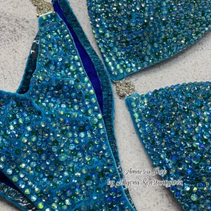 May include: A turquoise blue bikini top and bottom with crystal embellishments. The bikini is made of a soft, stretchy fabric and has a high-cut leg. The bikini is decorated with thousands of tiny crystals that sparkle in the light. The bikini is designed for competition and is perfect for a woman who wants to look her best on stage.