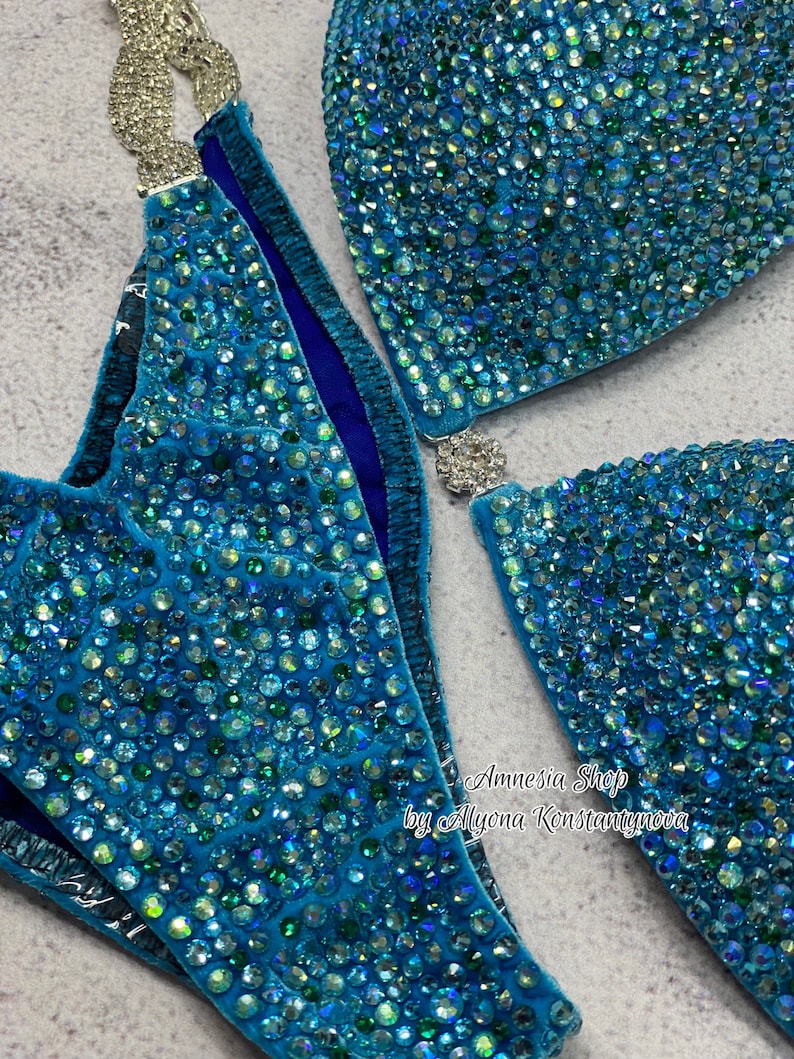 May include: A turquoise blue bikini top and bottom with crystal embellishments. The bikini is made of a soft, stretchy fabric and has a high-cut leg. The bikini is decorated with thousands of tiny crystals that sparkle in the light. The bikini is designed for competition and is perfect for a woman who wants to look her best on stage.