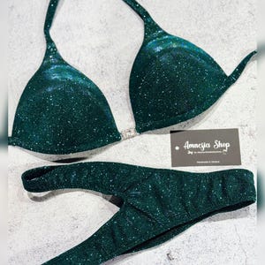 Puede incluir: Un top y una parte inferior de bikini verde oscuro con brillo. La parte superior tiene una correa de estilo halter y la parte inferior es de estilo de corte alto. El bikini está hecho a mano en Ucrania. La etiqueta dice "Amnesia Shop".