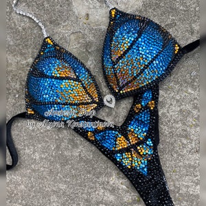 Op de afbeelding: Een zwarte bikini met een blauw en gouden vlinderontwerp gemaakt van strass. De bikini ligt op een grijze achtergrond. De tekst "Amnesia Shop by Alyona Konstantinova" staat op de bikini.