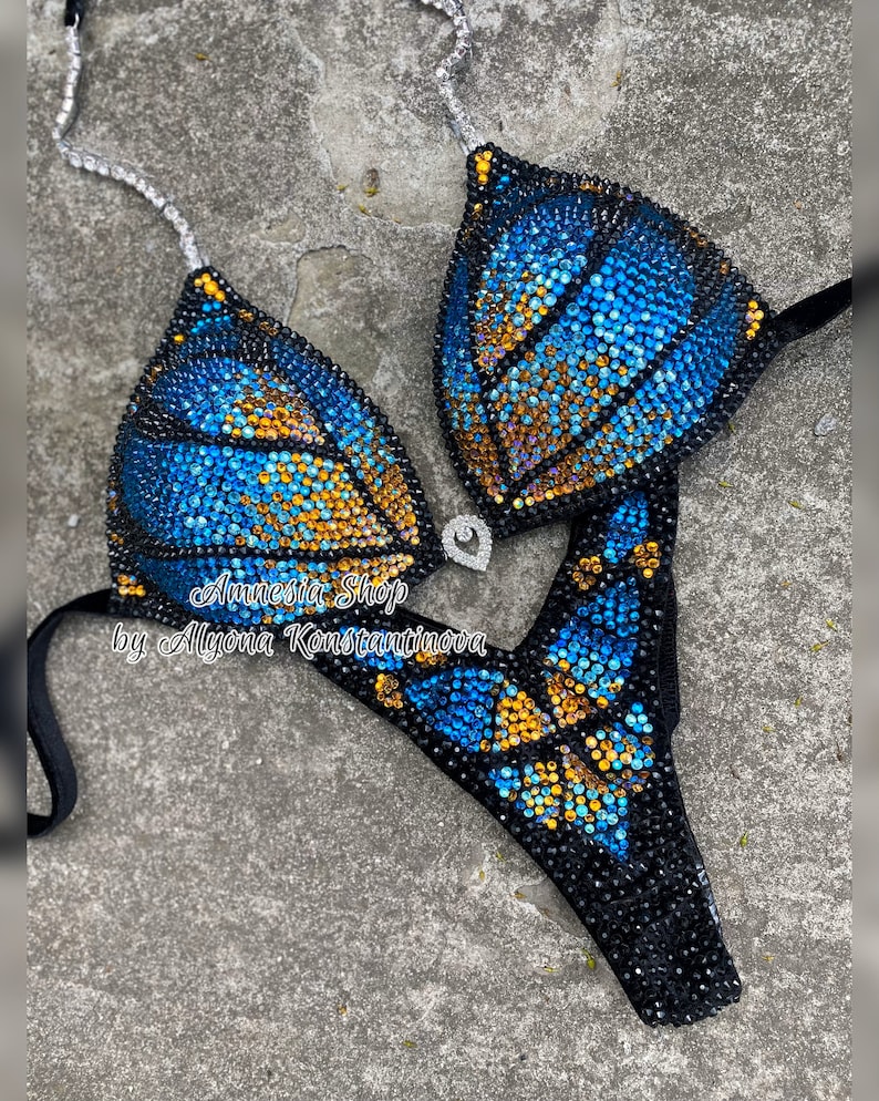 Op de afbeelding: Een zwarte bikini met een blauw en gouden vlinderontwerp gemaakt van strass. De bikini ligt op een grijze achtergrond. De tekst "Amnesia Shop by Alyona Konstantinova" staat op de bikini.