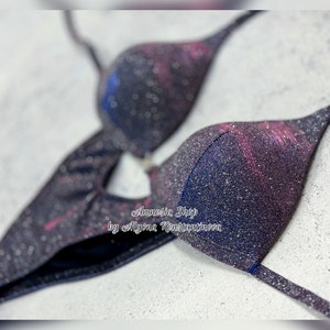 May include: A black and pink glittery bikini top with a galaxy print. The top has a halter style strap and a clasp closure in the back. The text "Amnesia Shop by Alyona Konstantinova" is printed on the top.