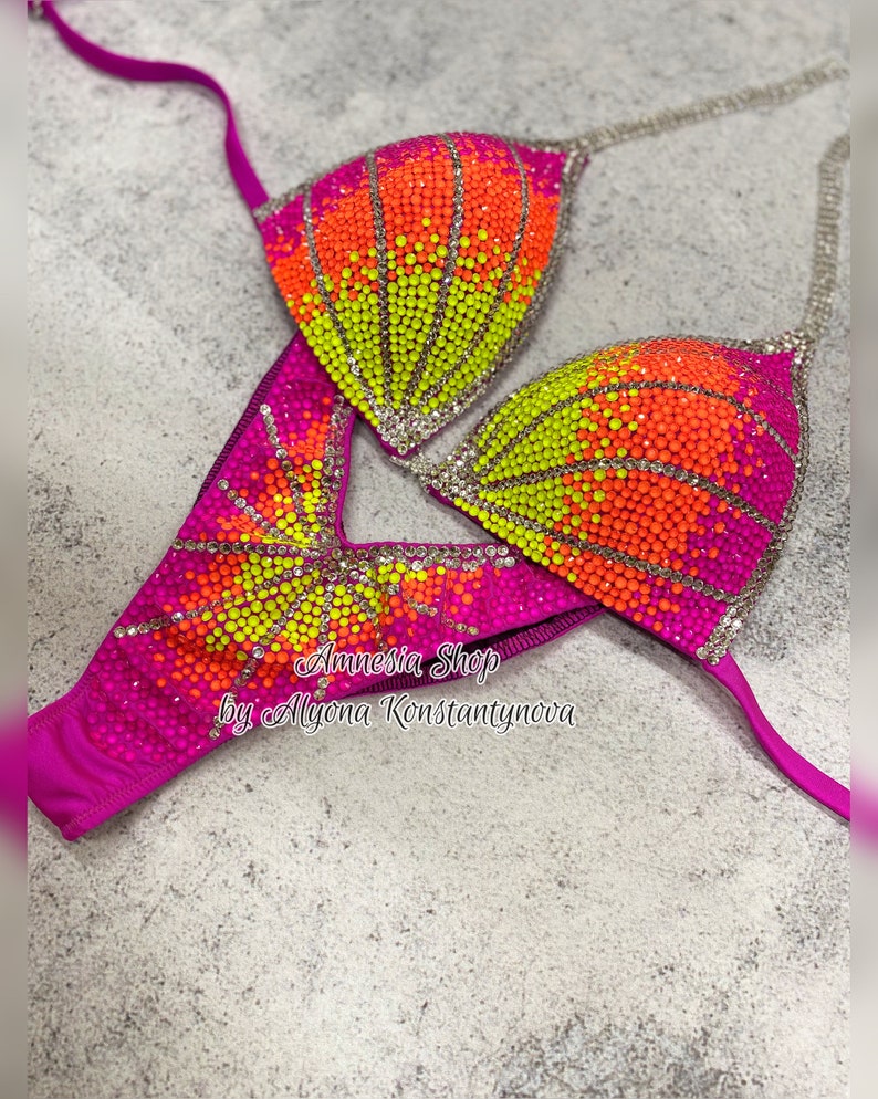 May include: A pink, yellow, and orange sequined bikini top and bottom with rhinestones. The bikini is designed with a sunburst pattern. The text "Amnesia Shop by Alyona Konstantynova" is printed on the bottom.