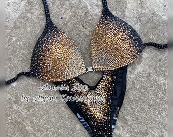 May include: A black and gold sequined bikini competition suit. The suit has a halter top and a high-cut bottom. The top is decorated with a gradient of black, gold, and silver sequins. The bottom is decorated with black and gold sequins.