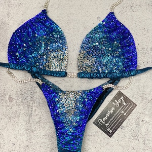 May include: A blue and silver sequined bikini with a crystal-embellished strap. The bikini is on a gray background with a business card that reads "Amnesia Shop by Ayana Konstantinova".