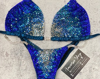 May include: A blue and silver sequined bikini with a crystal-embellished strap. The bikini is on a gray background with a business card that reads "Amnesia Shop by Ayana Konstantinova".