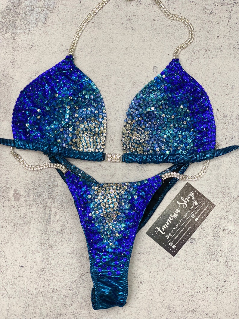 May include: A blue and silver sequined bikini with a crystal-embellished strap. The bikini is on a gray background with a business card that reads "Amnesia Shop by Ayana Konstantinova".