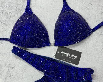 May include: A royal blue bikini with crystal embellishments. The bikini top has a halter style with a triangle cup. The bikini bottom is a high-cut style with a V-shaped front. The bikini is handmade in Ukraine. The tag reads "Amnesia Shop by Amnesia Underwear".