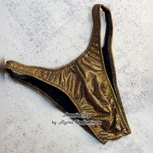 May include: Gold metallic swimsuit bottom with a black lining. The swimsuit has a low-cut leg opening and a high-cut back. The fabric has a shimmery finish. The text "Amnesia Shop by Alyona Konstantinova" is printed on the fabric.