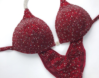 May include: A red, sequined, competition bikini with a silver strap design. The bikini is fully covered in small, red rhinestones.