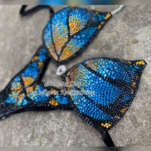 Op de afbeelding: Een zwarte bikini top met een vlinderontwerp gemaakt van blauwe en gouden strass steentjes. De tekst "Amnesia Shop by Alyona Konstantinova" is zichtbaar op de bikini top.
