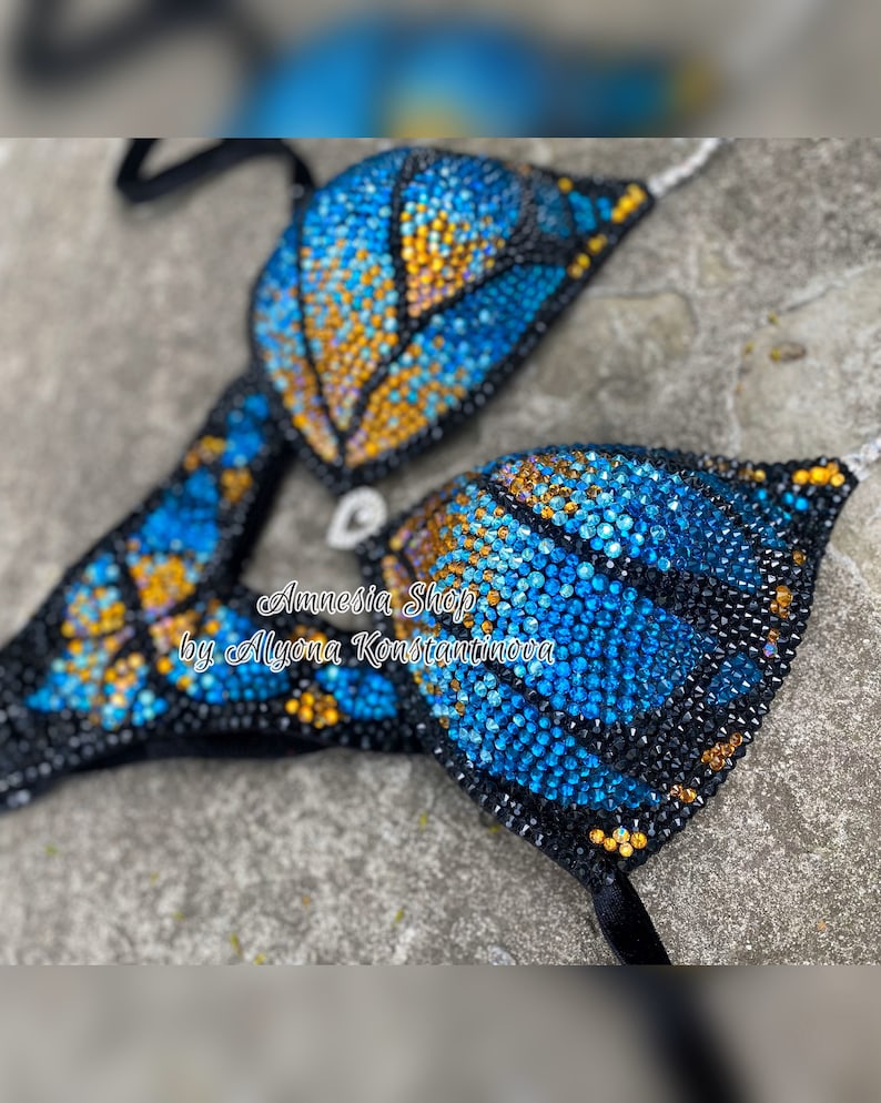 Op de afbeelding: Een zwarte bikini top met een vlinderontwerp gemaakt van blauwe en gouden strass steentjes. De tekst "Amnesia Shop by Alyona Konstantinova" is zichtbaar op de bikini top.