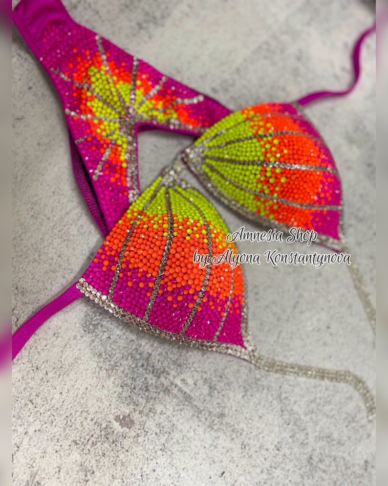 May include: A pink and green bikini top with rhinestones in a gradient pattern. The top is decorated with orange, yellow, and green rhinestones. The text "Amnesia Shop by Alyona Konstantynova" is visible on the top.