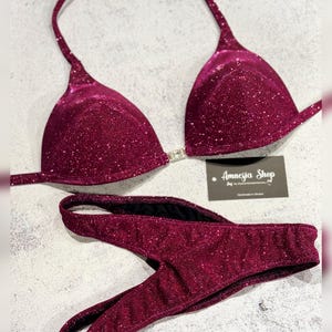 Puede incluir: Un bikini burdeos con un acabado brillante. La parte superior es de estilo triangular con una tira halter y la parte inferior es un corte en V. El bikini está hecho a mano en Ucrania por Amnesia Shop.