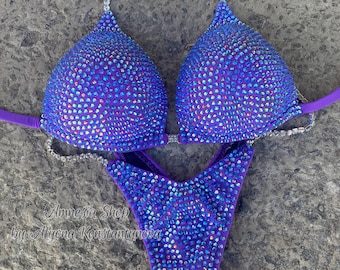 May include: Purple competition bikini with iridescent rhinestones. The bikini top has a halter style strap and the bottom has a V-shaped cut. The bikini is decorated with rhinestones.
