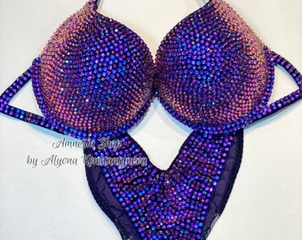 May include: Purple and pink crystal embellished competition bikini with a V-shaped bottom and a halter top. The bikini is designed by Amnesia Shop by Alyona Konstantynova.