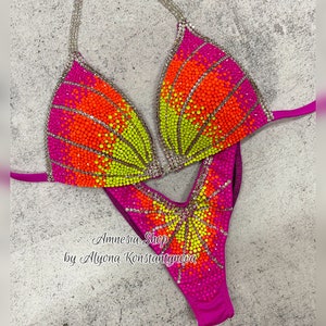 May include: A pink, orange, and yellow sequined competition bikini with a V-shaped neckline and a high-cut leg. The bikini is decorated with rhinestones and has a "Amnesia Shop by Alyona Konstantynova" label.
