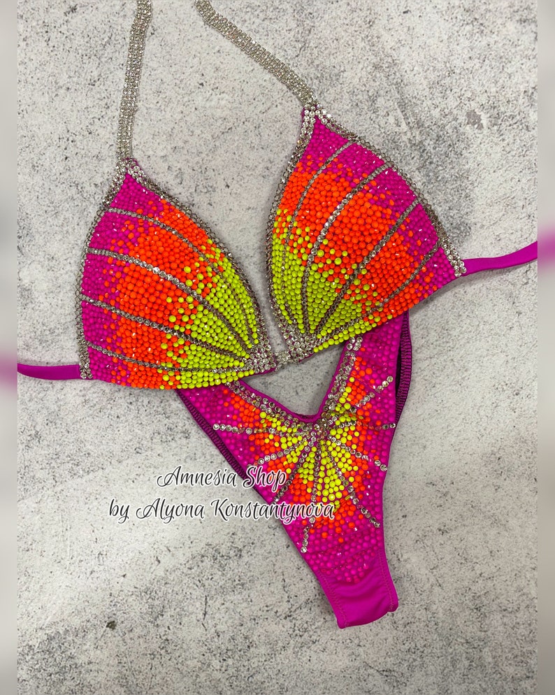 May include: A pink, orange, and yellow sequined competition bikini with a V-shaped neckline and a high-cut leg. The bikini is decorated with rhinestones and has a "Amnesia Shop by Alyona Konstantynova" label.