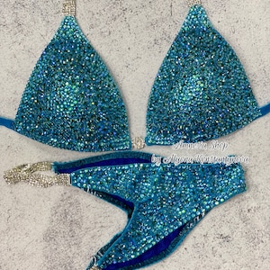 May include: A turquoise blue bikini competition suit with crystal embellishments and silver straps. The suit has a triangle top and a cheeky bottom. The text "Amnesia Shop by Alyona Konstantynova" is printed on the bottom.
