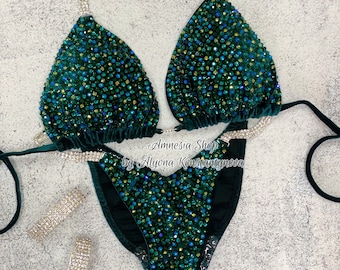 May include: A dark green velvet bikini with crystal embellishments. The bikini top has a triangle shape and the bottom is a high-cut style. The bikini is decorated with green, blue, and gold crystals. The text "Amnesia Shop by Alyona Konstantynova" is printed on the bottom of the bikini.