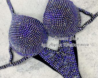 Könnte beinhalten: Lilafarbener und schwarzer Wettkampf-Bikini mit Kristallverzierungen. Der Bikini ist mit einem Neckholder-Oberteil und einem hochgeschnittenen Unterteil designt. Der Text "Amnesia Shop by Alyona Konstantinova" ist auf dem Bikini aufgedruckt.