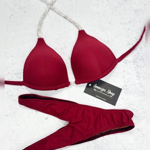 May include: Burgundy bikini top and bottom with a crystal-embellished strap. The bikini top is triangle shaped and the bottom is a high-cut style with a V-shaped opening at the front. The bikini is labelled "Amnesia Shop".