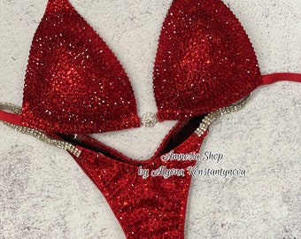 May include: Red and silver sequined bikini competition suit. The suit has a halter top and a high-cut bottom. The top is decorated with silver rhinestones and the bottom is decorated with red rhinestones. The suit is designed by Amnesia Shop by Allyonа Konstantynova.