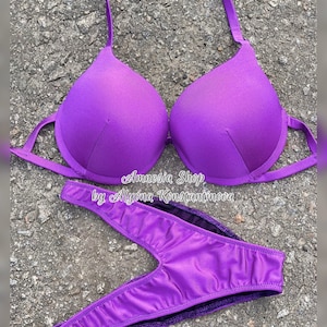 Puede incluir: Parte superior e inferior de bikini morado con un corte en V en la parte inferior. La parte superior del bikini tiene una correa de estilo halter y la parte inferior tiene una pierna de corte alto. El texto "Amnesia Shop by Alyona Konstantinova" está impreso en la parte inferior.