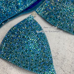 May include: A turquoise blue bikini top with crystal embellishments. The top is made of a stretchy fabric and has a triangle shape. The bikini top is decorated with iridescent crystals that sparkle in the light. The text "Amnesia Shop by Alyona Konstantynova" is printed on the top.