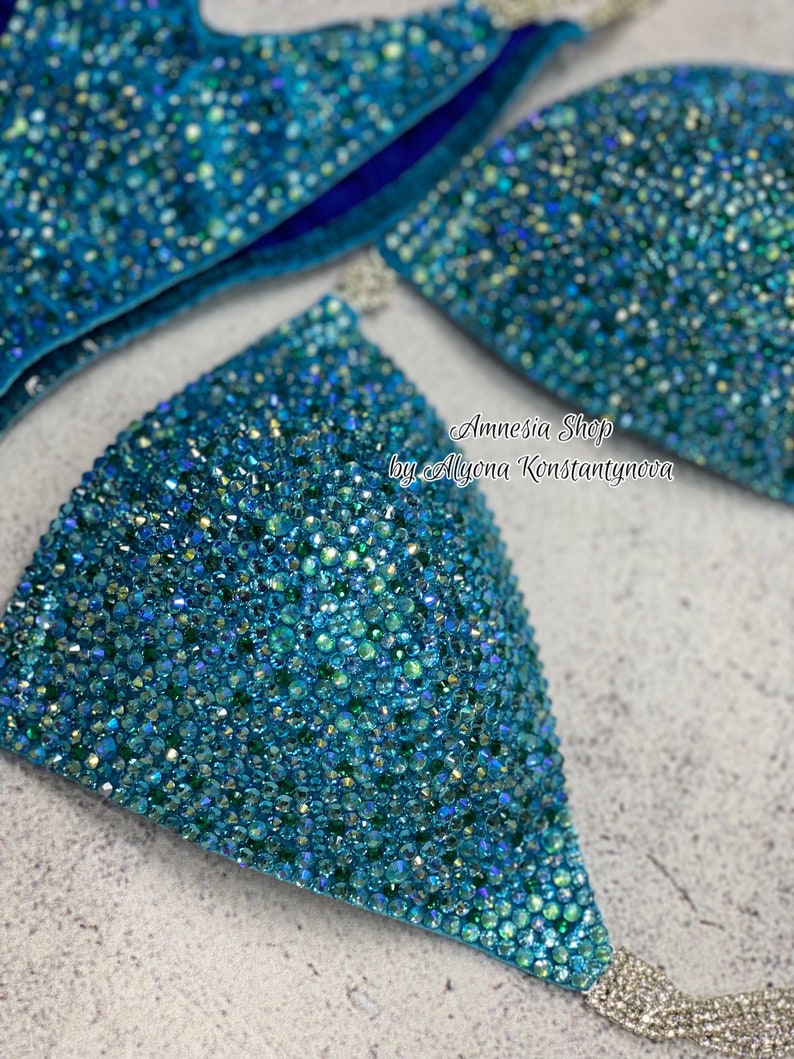 May include: A turquoise blue bikini top with crystal embellishments. The top is made of a stretchy fabric and has a triangle shape. The bikini top is decorated with iridescent crystals that sparkle in the light. The text "Amnesia Shop by Alyona Konstantynova" is printed on the top.