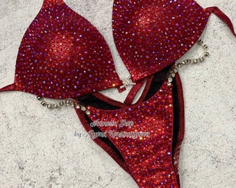 Op de afbeelding: Rode, glinsterende wedstrijd bikini met kristallen versieringen en een zwarte mesh voering. De bikini is ontworpen voor figuurwedstrijden en heeft een haltertop en een hoog uitgesneden onderkant. De tekst "Amnesia Shop by Alyona Konstantynova" is gedrukt op de onderkant.