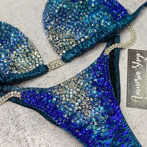 May include: A teal blue bikini with a gradient of blue rhinestones. The bikini top has a triangle shape and the bottom is a high-cut style. The bikini is decorated with a variety of blue rhinestones, including clear, light blue, and dark blue.