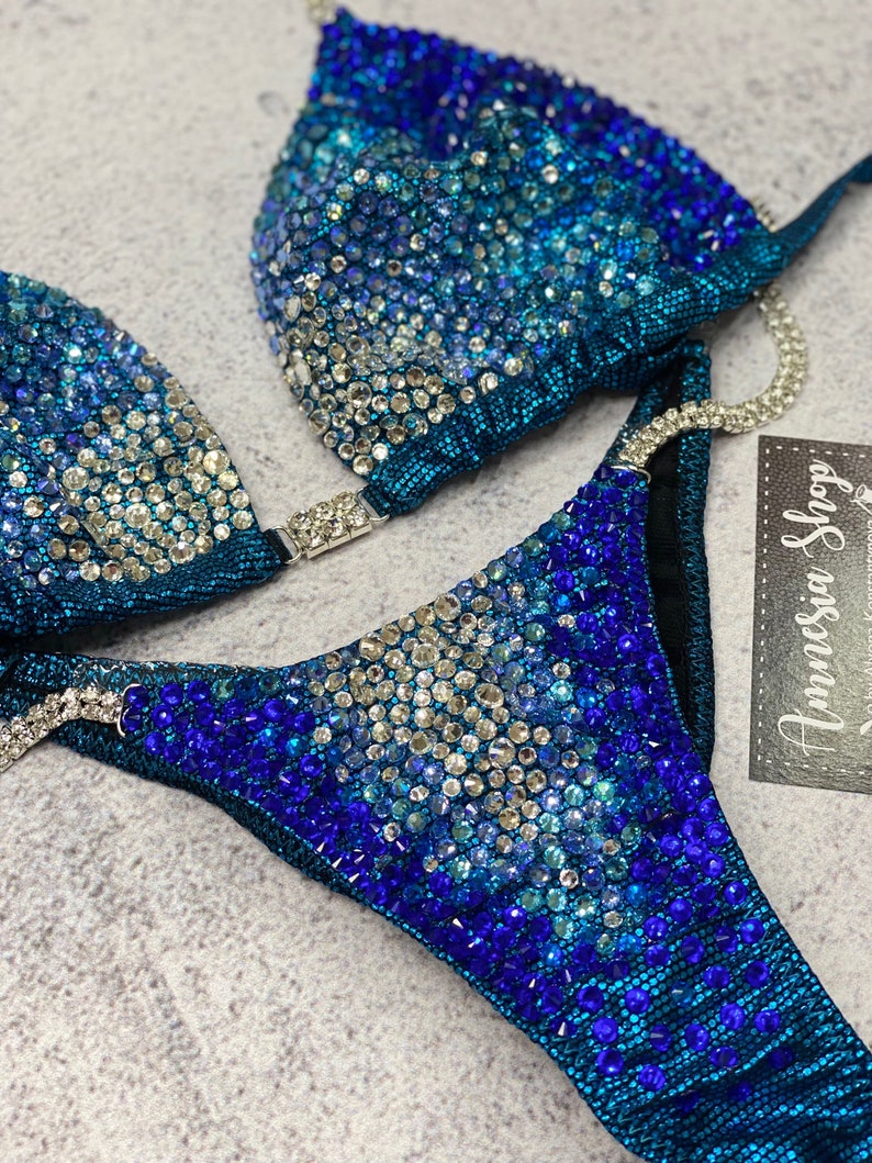 May include: A teal blue bikini with a gradient of blue rhinestones. The bikini top has a triangle shape and the bottom is a high-cut style. The bikini is decorated with a variety of blue rhinestones, including clear, light blue, and dark blue.